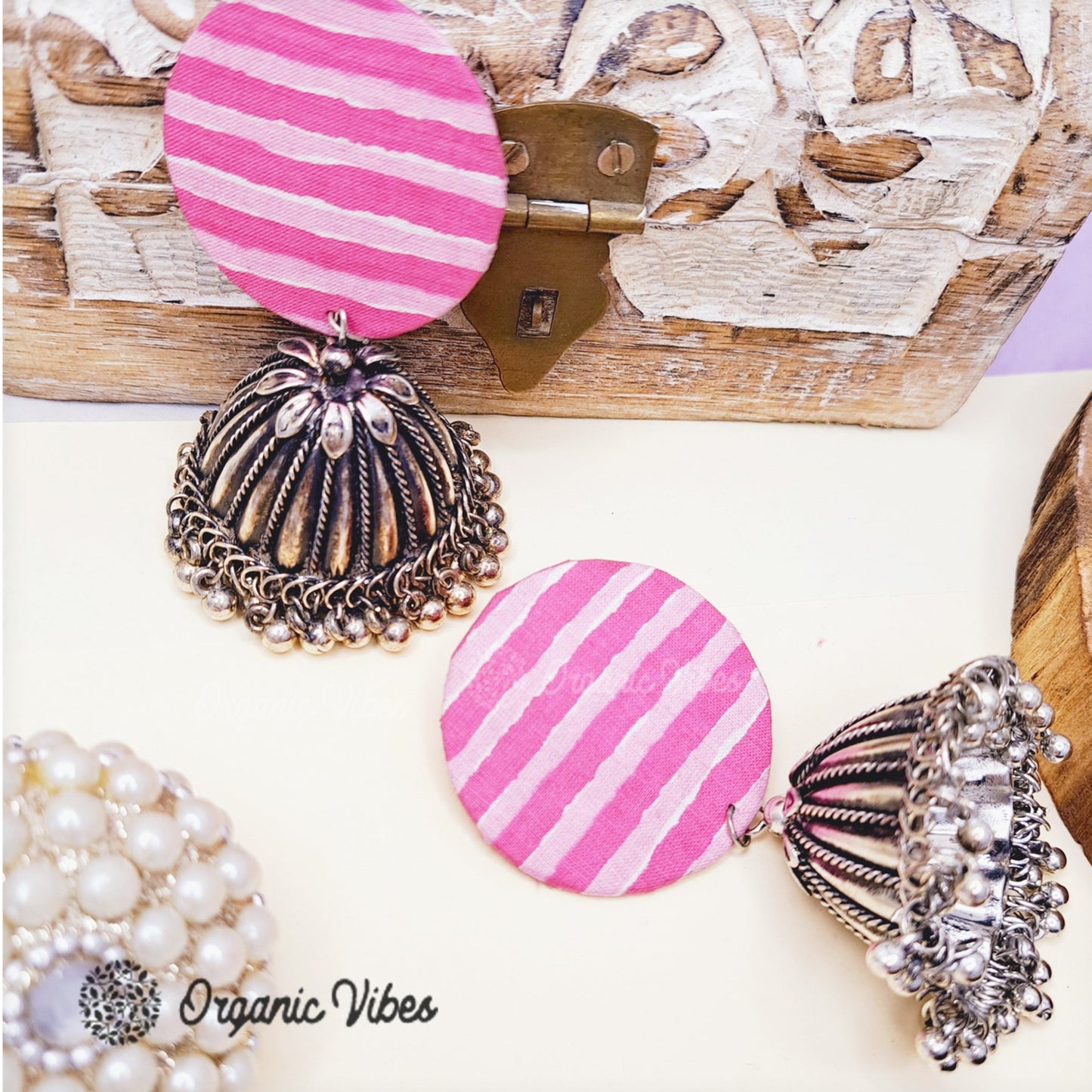 A pair of handmade fabric earrings with a pink and white striped pattern and silver jhumka design, displayed against a wooden background with decorative elements.