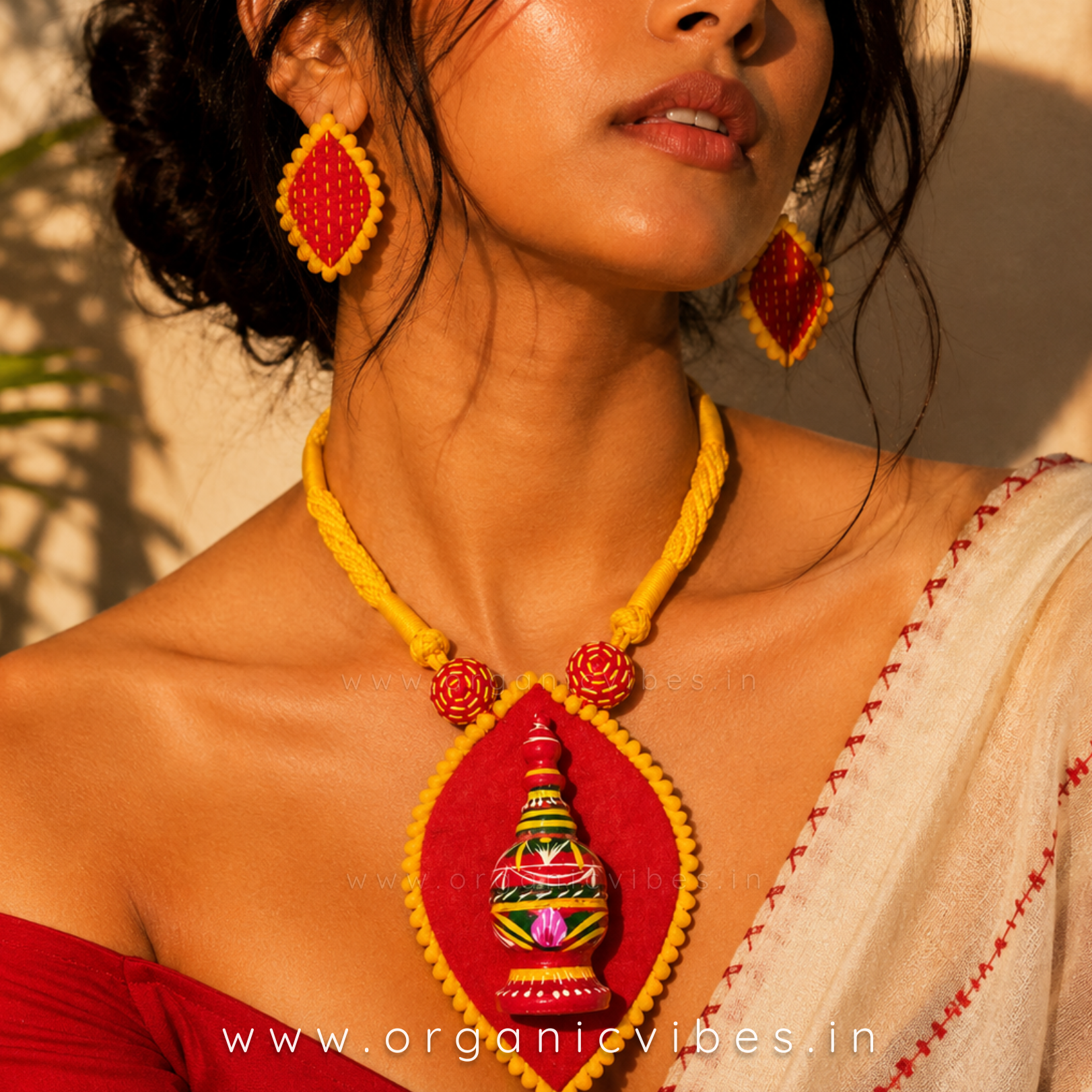 A woman wearing a handmade red and yellow beaded necklace and matching teardrop earrings by Organic Vibes, styled with a cream saree against a warm sunlit wall.