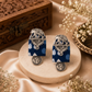 A pair of ornate silver-toned earrings with elephant motifs and blue patterned rectangular drops, displayed on a round beige stone pedestal with decorative dried flowers and a carved wooden box in the background.