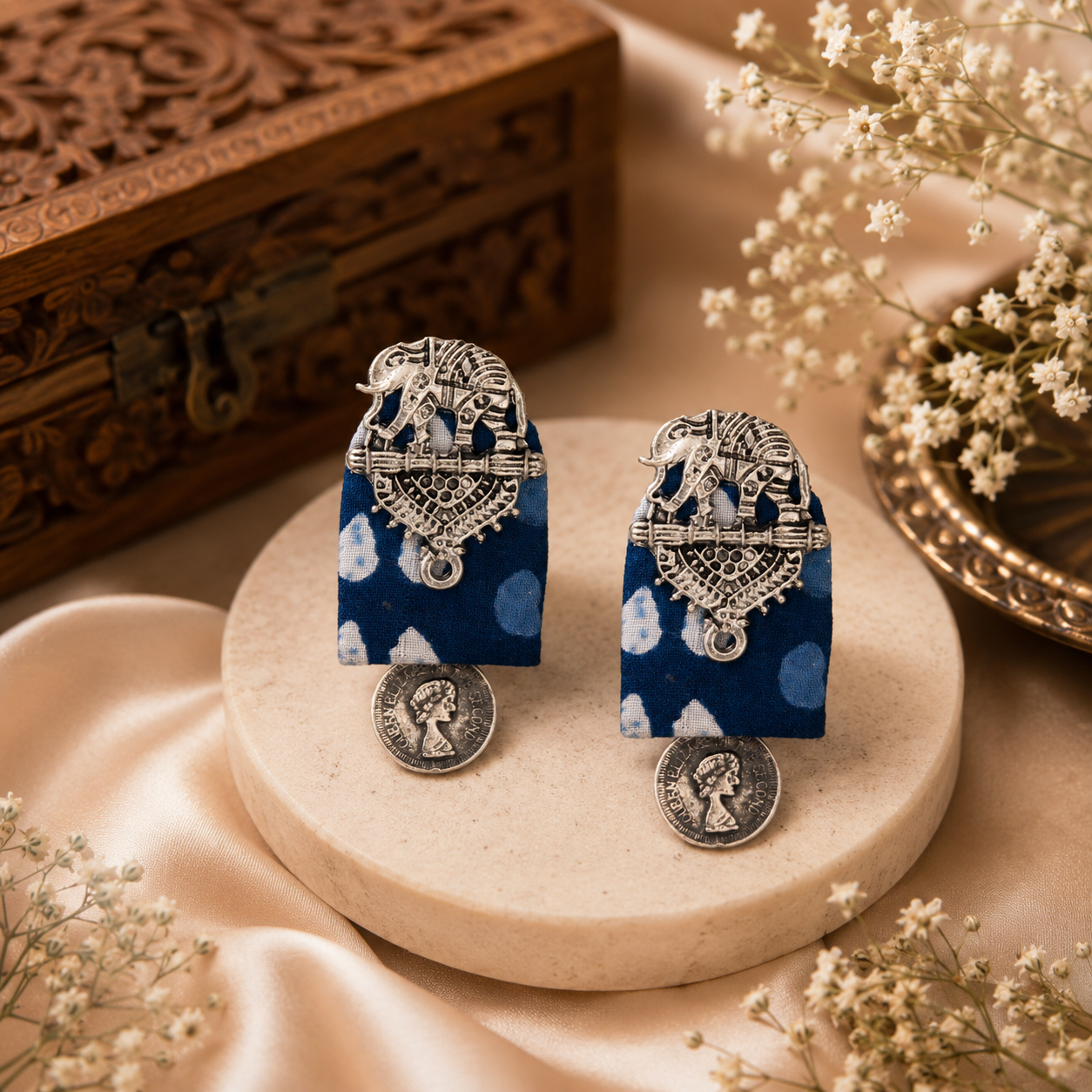 A pair of ornate silver-toned earrings with elephant motifs and blue patterned rectangular drops, displayed on a round beige stone pedestal with decorative dried flowers and a carved wooden box in the background.