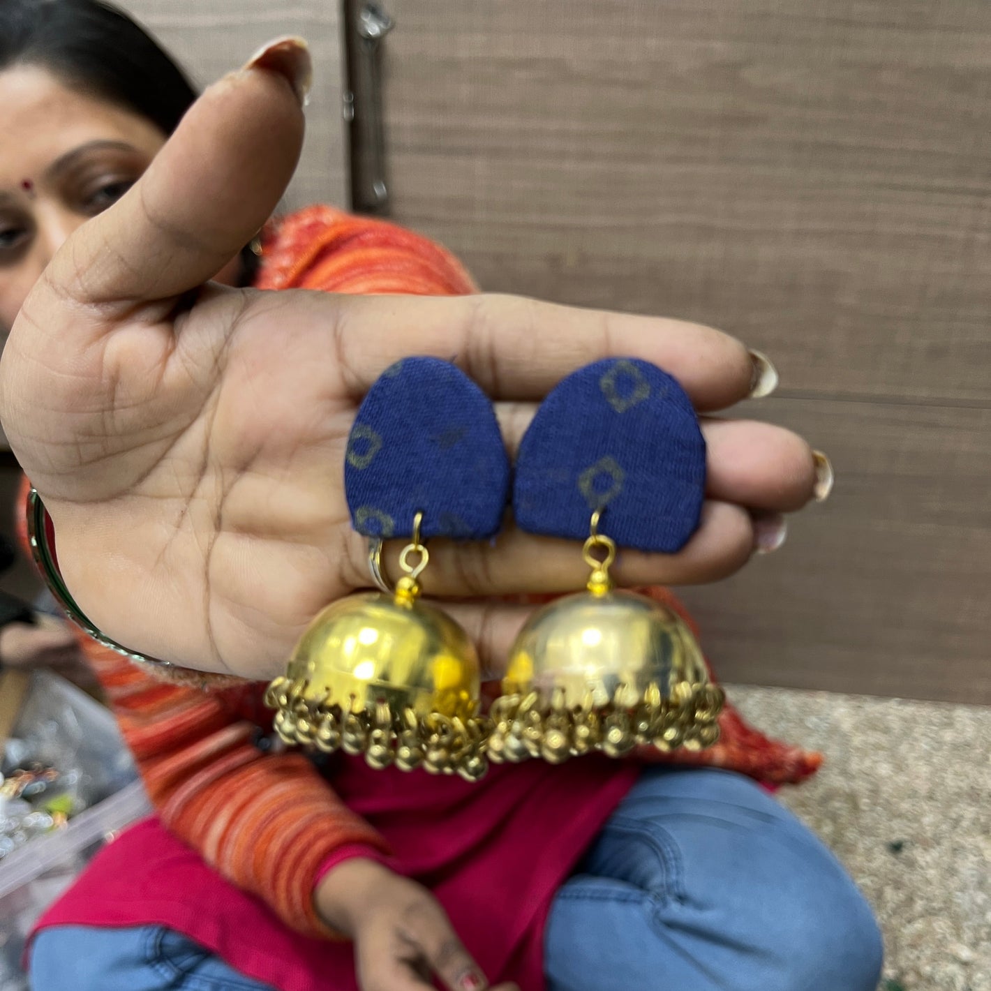 A person holds up a pair of handmade earrings with blue fabric studs and shiny gold bell-shaped dangling charms, with a woman sitting in the background.