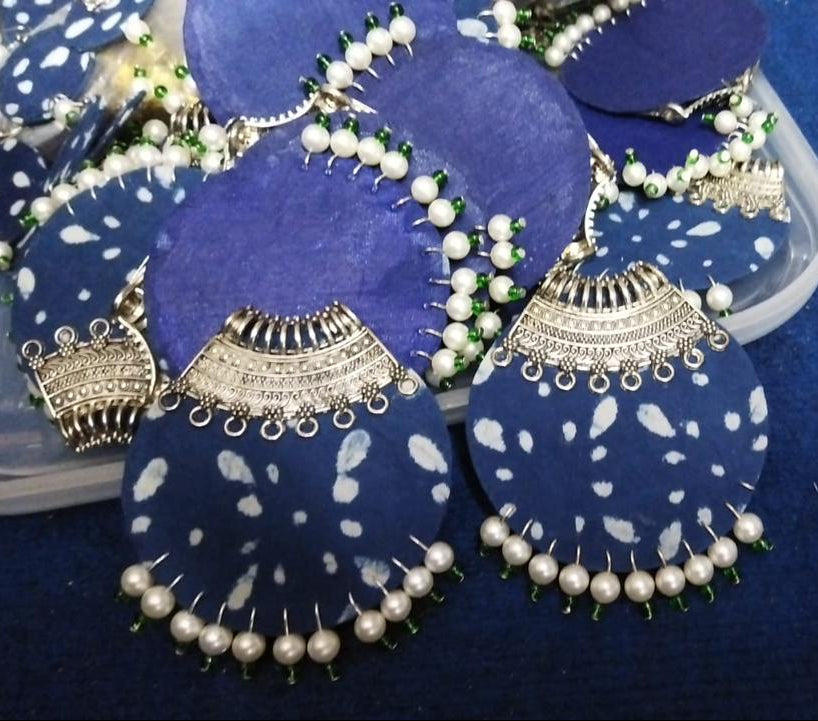 A close-up of a cluster of handmade blue and white patterned earrings with silver-toned metal borders and white pearl bead fringe, arranged on a blue surface.
