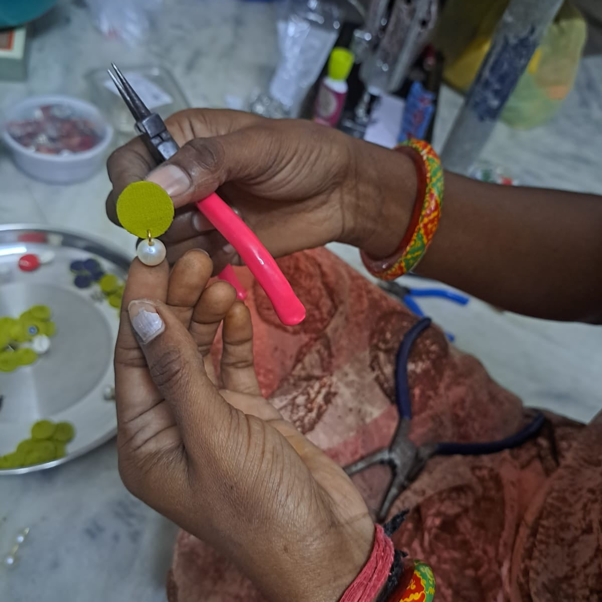 A close-up of a person's hands holding a small craft tool and a yellow circular embellishment with a pearl bead, working in a jewelry-making or craft workspace.